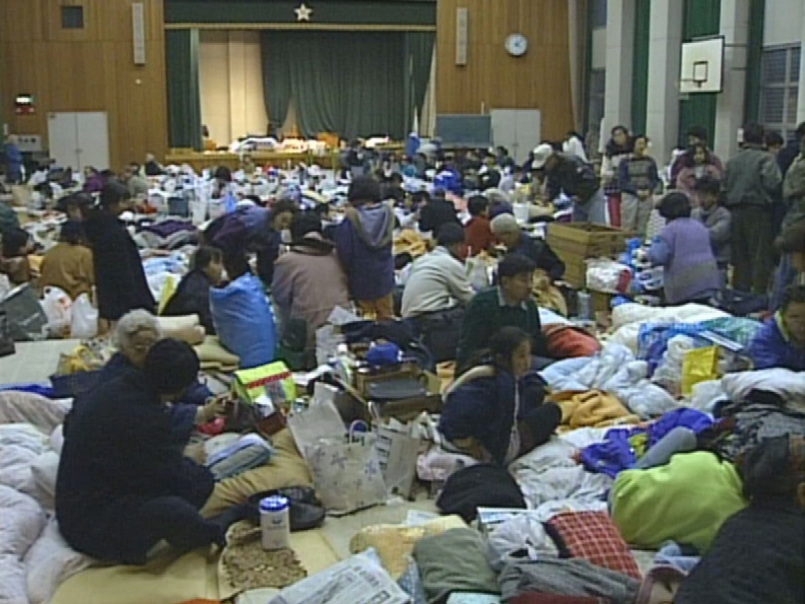 体育館に設けられた避難所で人々が生活物資とともに過ごしている様子。